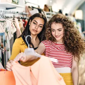 Two female teenager friends standing inside in the shop, holding and looking at dress.