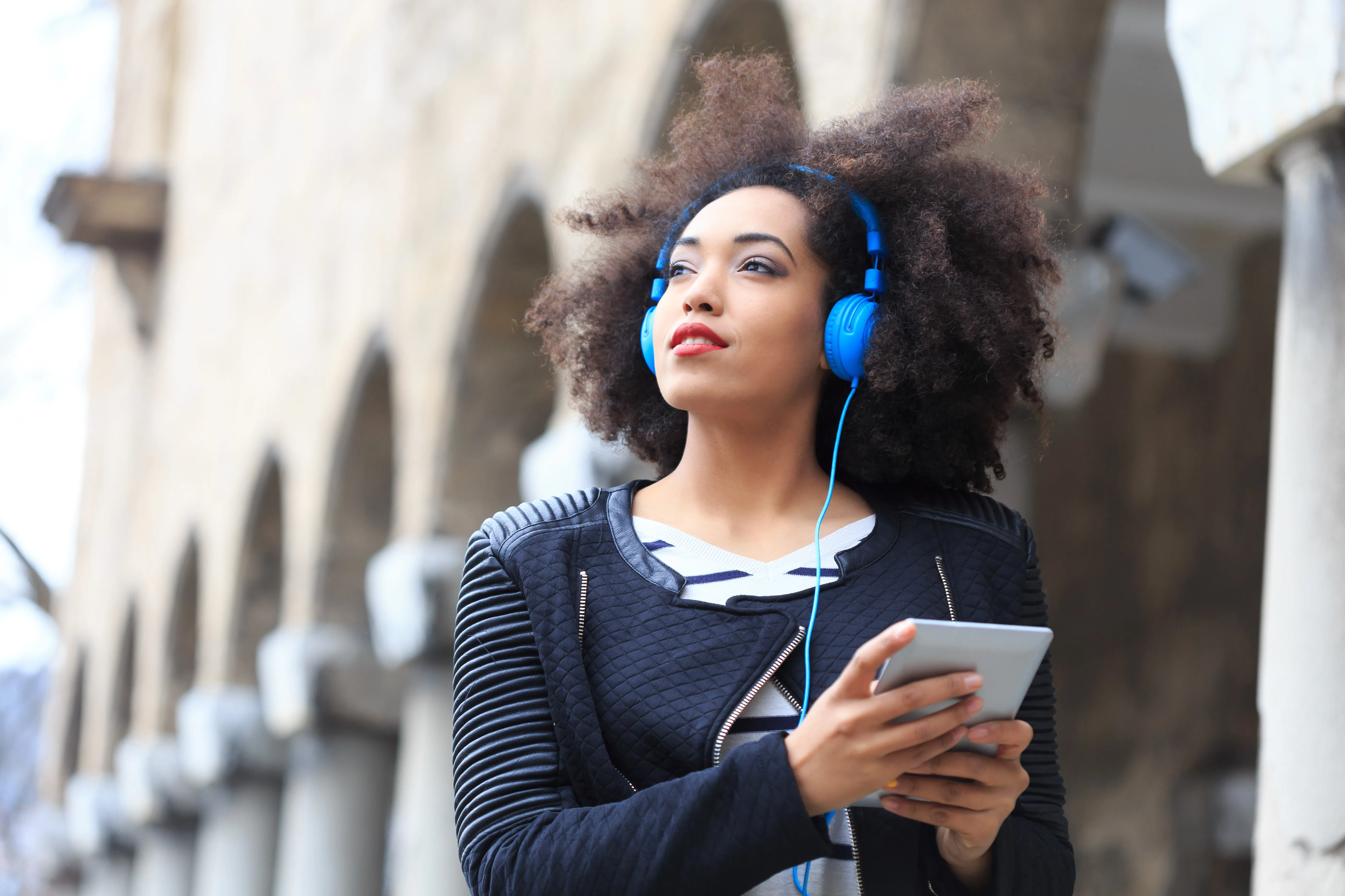 woman with curly afro looking away from the camera while standing outside with blue head phones around her head that are connected to a tablet