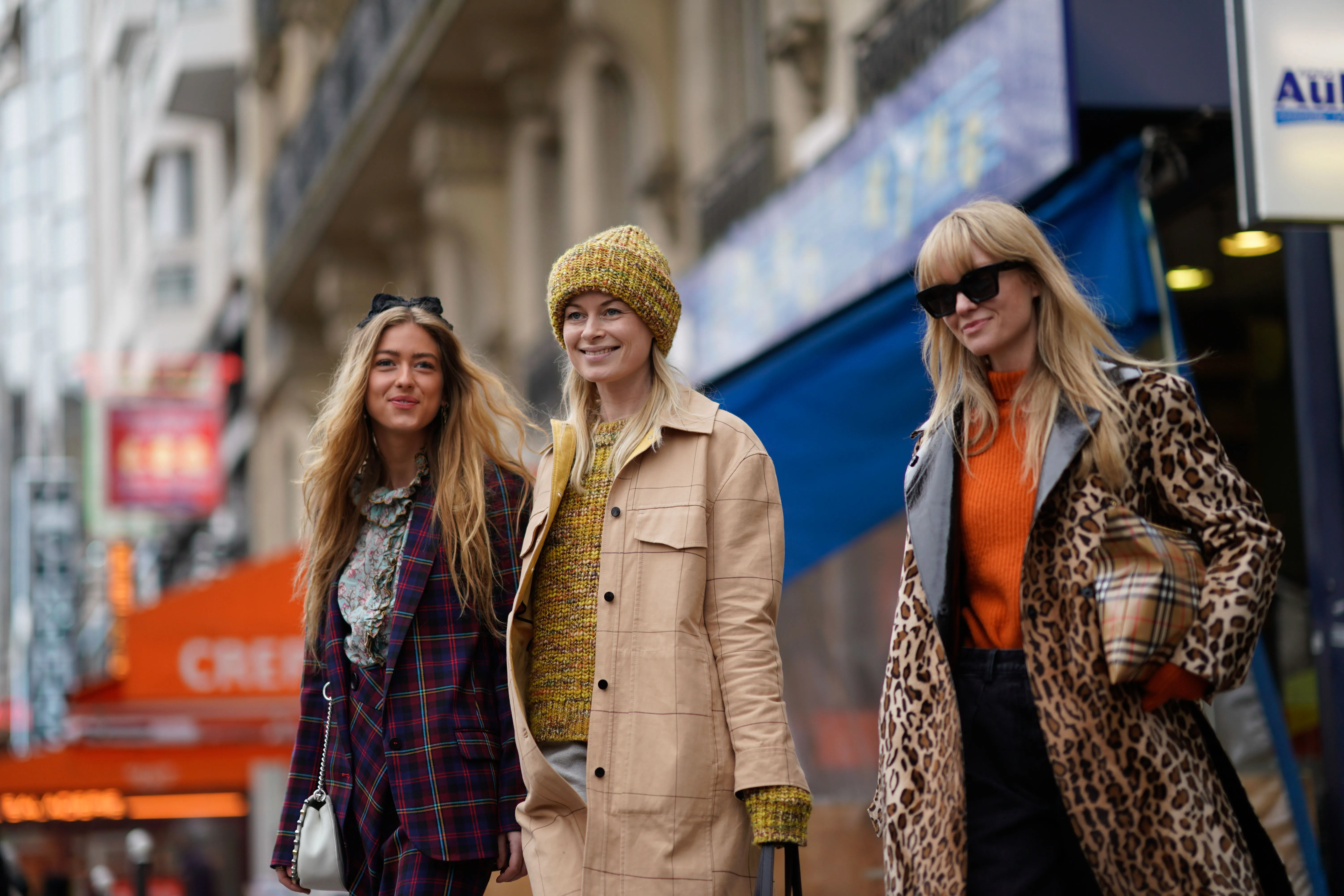 three women walking wearng coats. One woman is wearing a khaki coat, another is wearing a plaid blazer coat another is wearing a leopard print coat. All are smiling.
