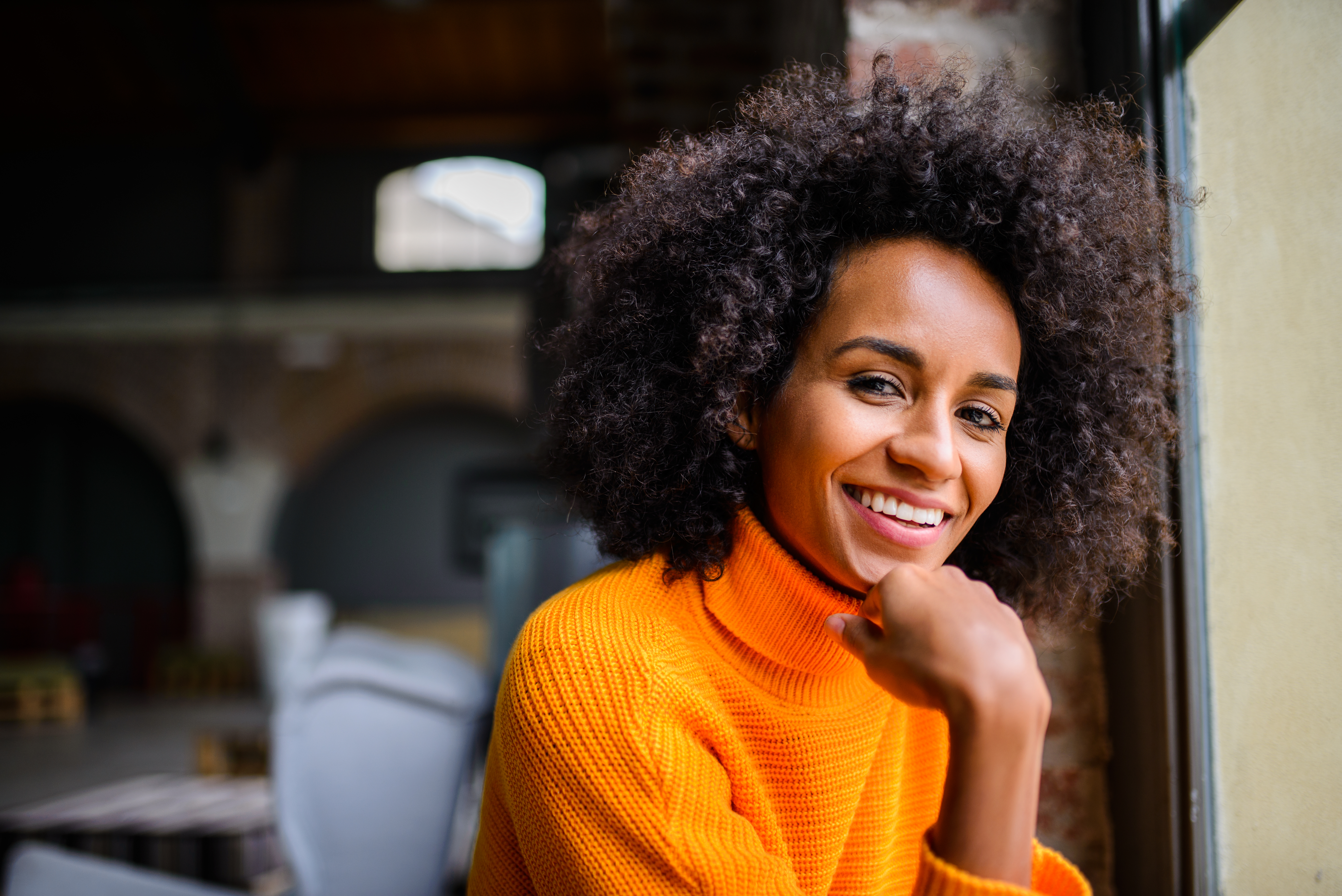 woman with big curly hair smiling while sitting down wearing a bright orange sweater