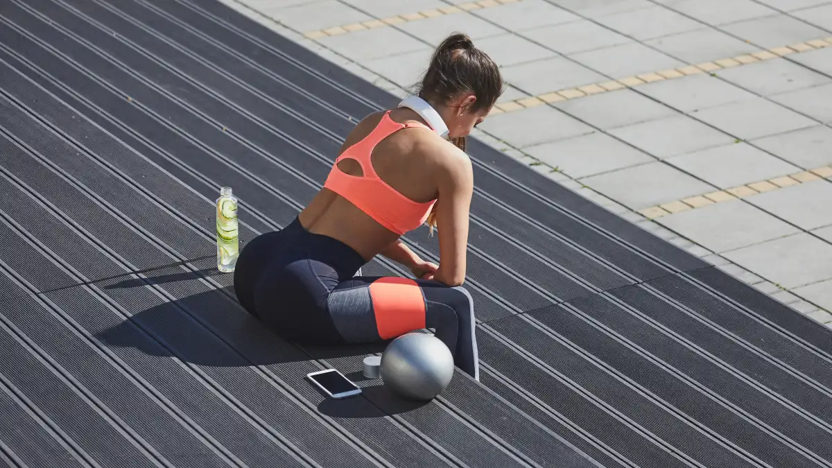 Woman sitting and taking a rest from exercising. Next to her fitness ball, smart phone and refreshing drink.