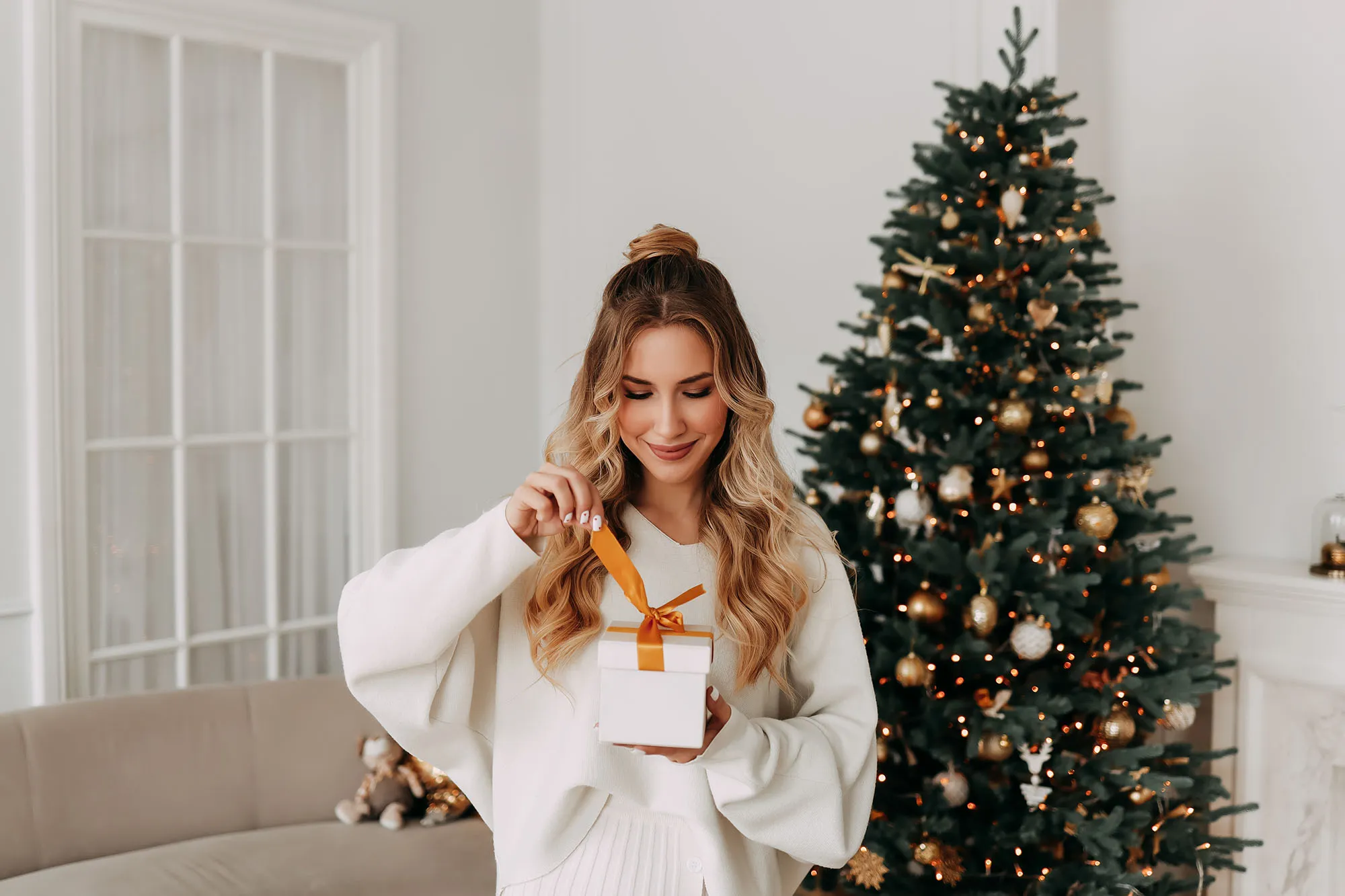 The concept of Christmas. Portrait of a smiling young woman with curly hair in a knitted sweater holding and opening a Christmas gift on the background of a Christmas tree in a bright interior on a holiday at home