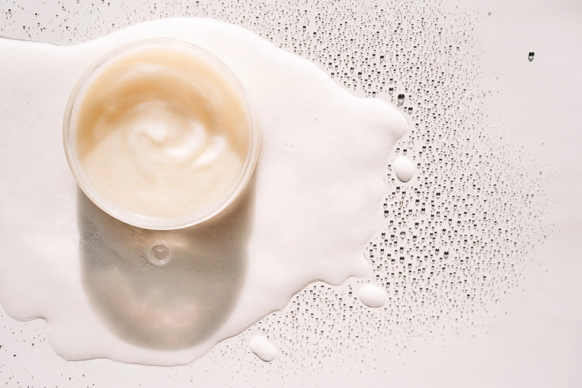 Glass jar with beige cream on white foam among many drops. Concept of body care and beauty. Copy space for your design. Flat lay style