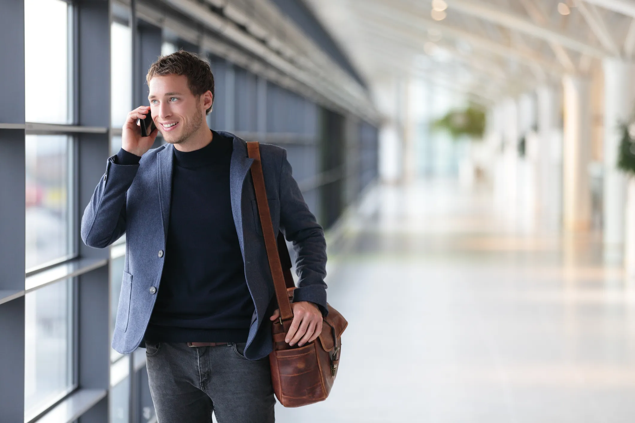 highly rated messenger bag Urban business man talking on smart phone traveling walking inside in airport. Casual young businessman wearing suit jacket and shoulder bag. Handsome male model in his 20s.