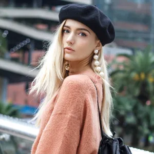 A beautiful young blonde woman dressed in a large warm pink sweater with a black beret and gorgeous earrings.