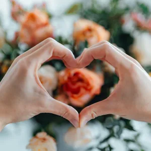 Woman forming a heart with hands against flower bouquet. Heart made with hands. Receiving flower bouquet delivery on Valentine’s day. Online dating concept. Long distance relationship. Dating anniversary.