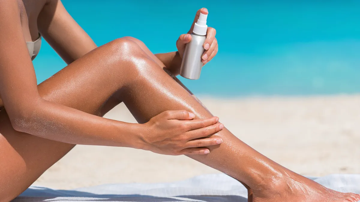 Sunscreen suntan lotion in spray bottle. Young woman in spraying tanning oil on her leg from bottle. Lady is massaging sunscreen lotion while sunbathing at beach. Female model during summer vacation.