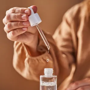 A girl in a beige dress holds a pipette with a drop of serum in her hands. Horizontal.