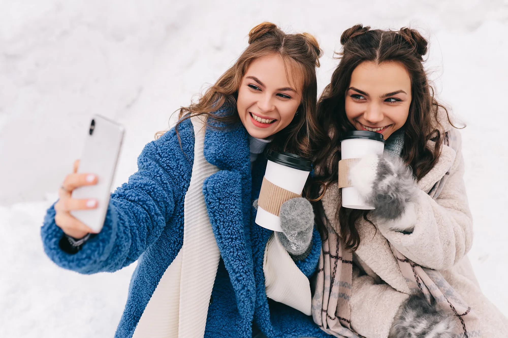 Two happy girls millennials teenagers friends are walking, laughing, taking selfies using wireless technology and a mobile phone, skating on a skating rink in a winter forest during the Christmas holidays. A couple of women in love spends time together in nature