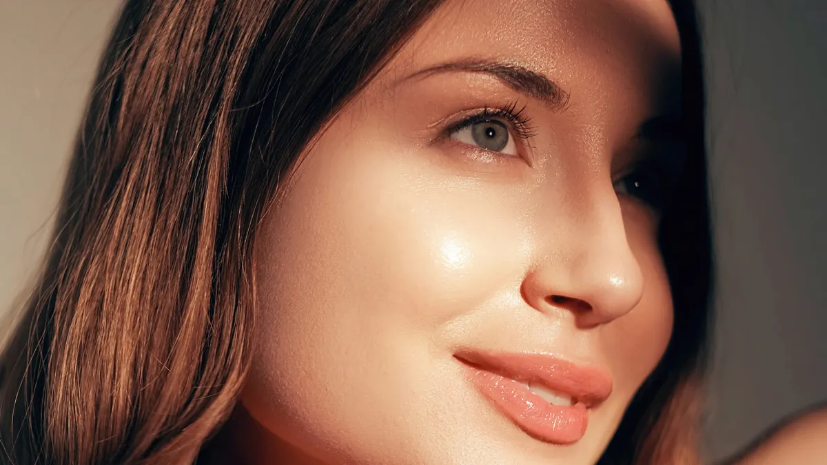 Beauty care. Radiant face. Moisturized skin. Cosmetology treatment. Pretty smiling young fresh glowing woman with glossy lip makeup enjoying sunshine isolated on beige.