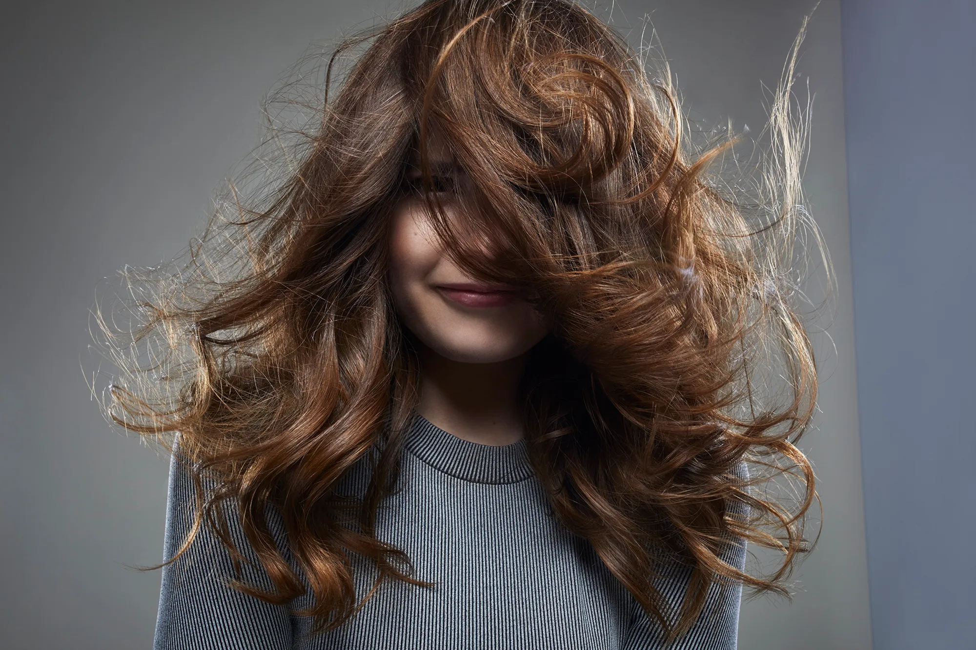 studio shot of a young female brunette in front of a gray background, wearing a gray outfit . her face half hidden behind her hair
