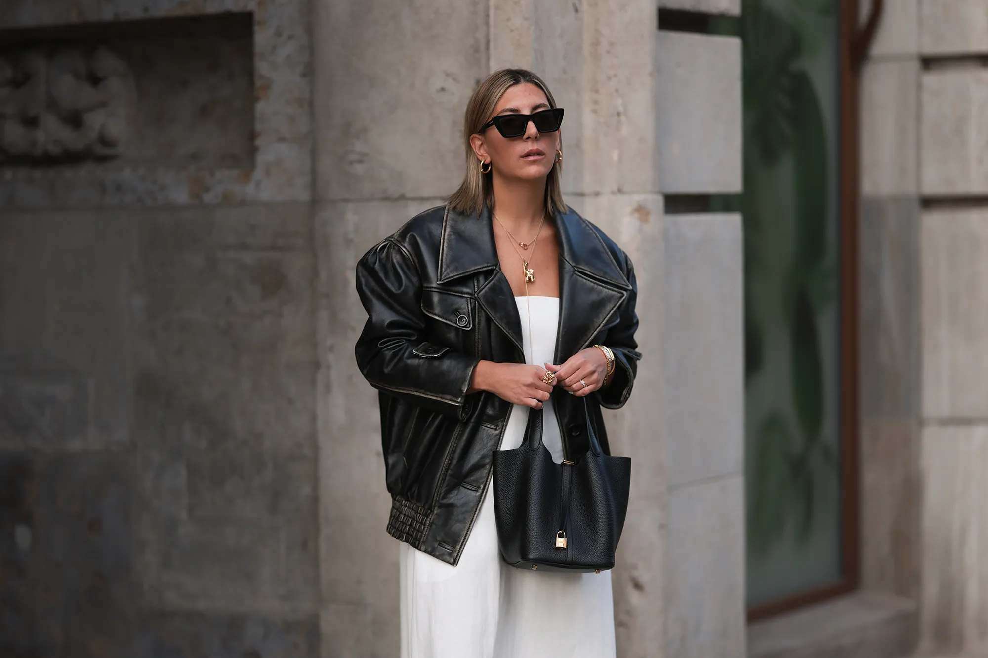 Aylin Koenig seen wearing Saint Laurent black sunglasses, Cartier Love gold yellow necklace, Ole Lynggaard elephant yellow gold necklace, Miu Miu black oversized leather jacket, By Aylin Koenig white bandeau long dress, Hermès Picotin black leather bag, Cartier yellow gold Juste Un Clou bracelet and Cartier yellow gold Love bracelet, on March 07, 2024 in Hamburg, Germany.