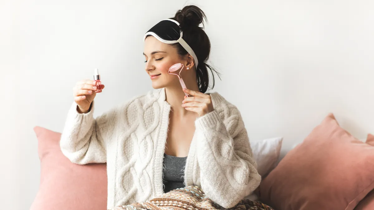 A girl with nice skin and her hair in a bun is sitting down comfortably leaning on pillows, legs covered with blanket. She is wearing a soft white fluffy cardigan and a facemask on her head. Beautician is holding up a small bottle of skincare oil and applying it as she's massaging her face with a special roller. It's very relaxing and she is smiling pleasantly. Self care concept.