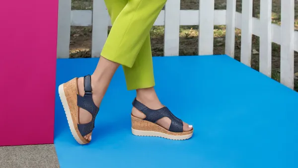 Outdoor scene featuring a woman modeling stylish navy blue wedge sandals, cork soles and perforated straps. stands on a bright blue floor, pink wall, wearing lime cropped pants, greenery background.