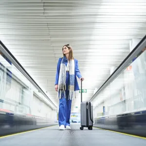Low angle view of cheerful woman standing in the airport moving walkway with her travel suitcase and looking away. Travel concept.