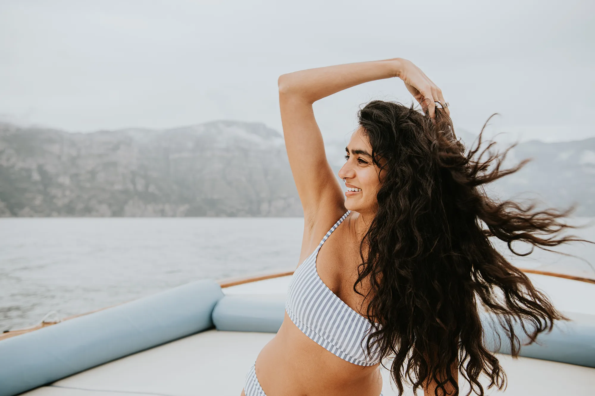 A beautiful young Indian woman enjoys a boat ride. She runs her fingers through her long, thick, wavy hair, which blows in the breeze. She looks away.