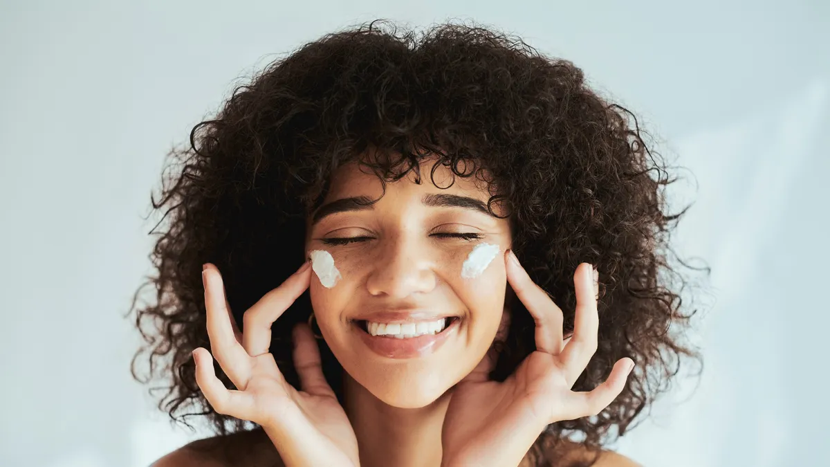 Moisturizer, cream on face in cosmetics, beauty or skincare promotion on white wall background. Facial, happy person or model smile with lotion, essential oil or sunscreen application