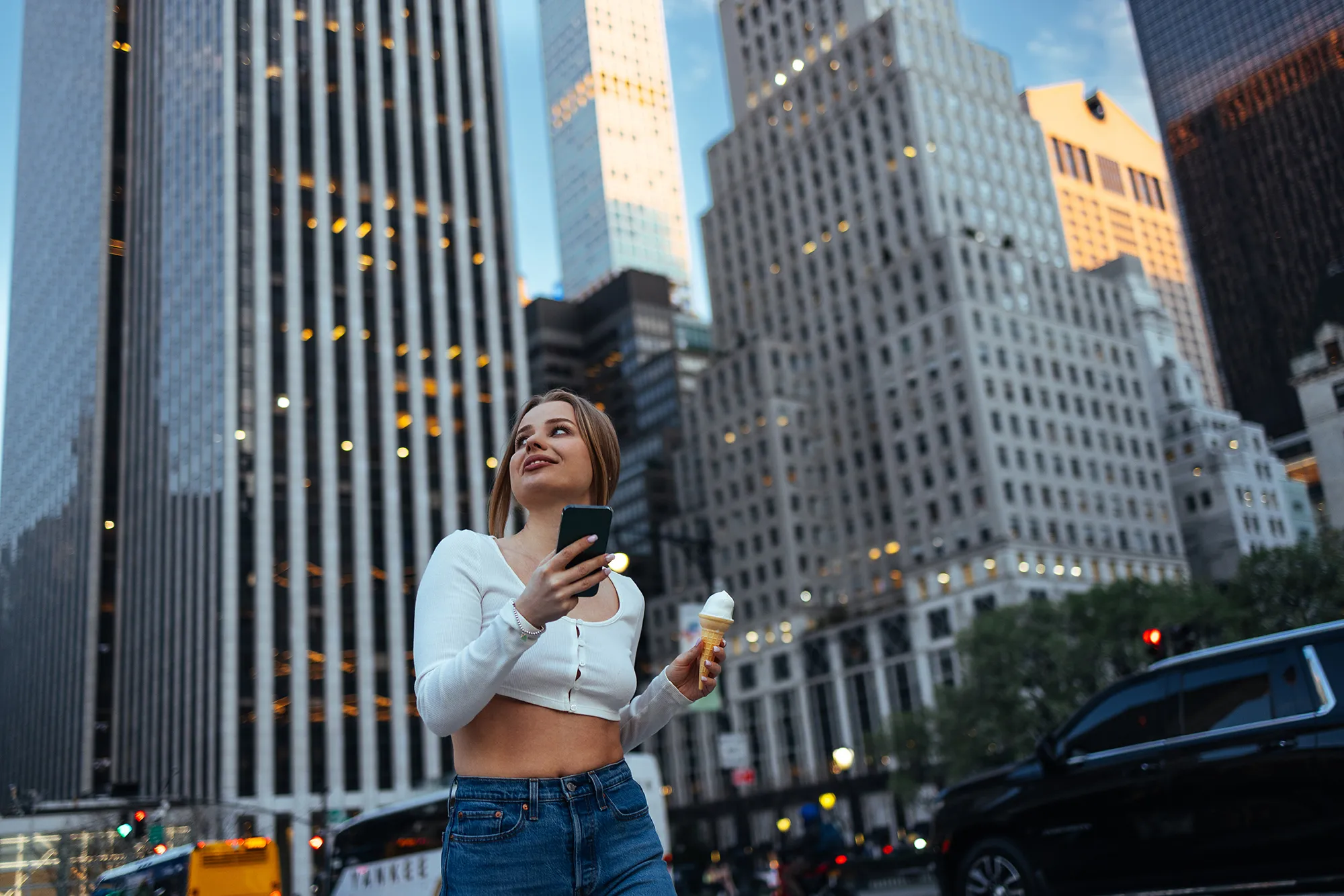 A woman gazes upward while holding her phone and ice cream cone, city buildings behind her