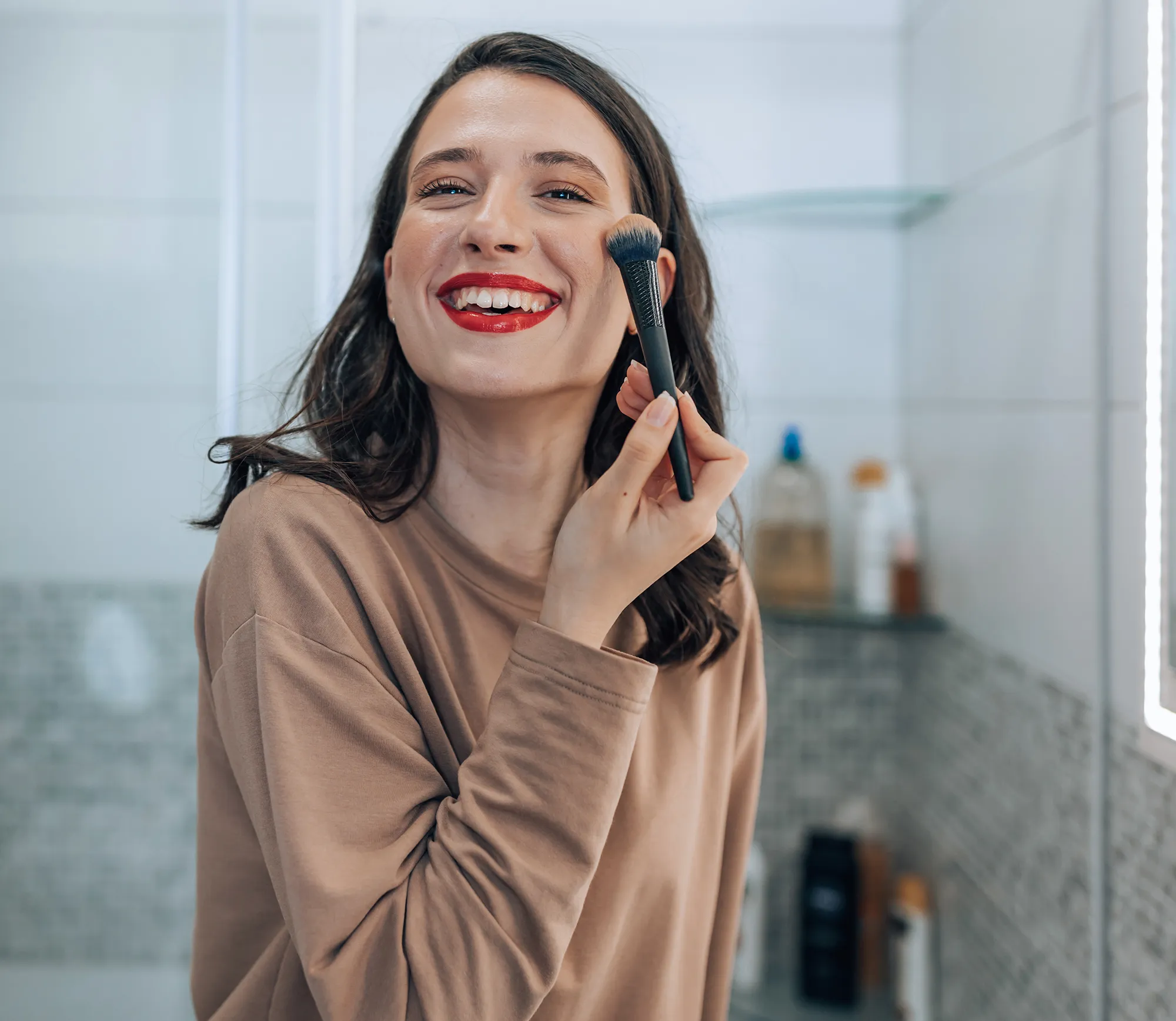 A young woman wearing red lipstick is looking at the camera with a big smile while applying blush to her cheeks.
