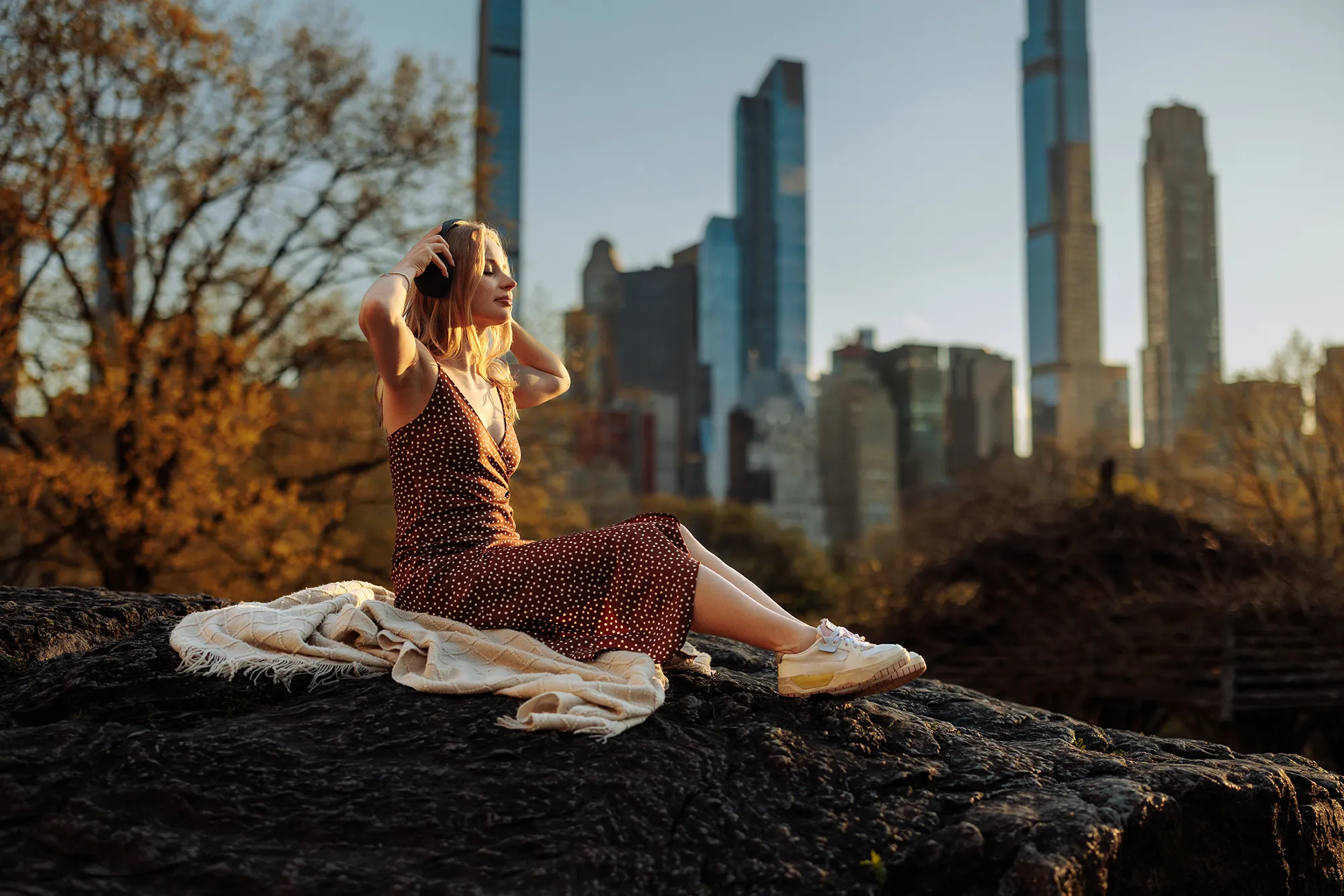 A woman sits serenely with a blanket, gazing at a city skyline from a vantage point, invoking a sense of peace despite the busy city backdrop
