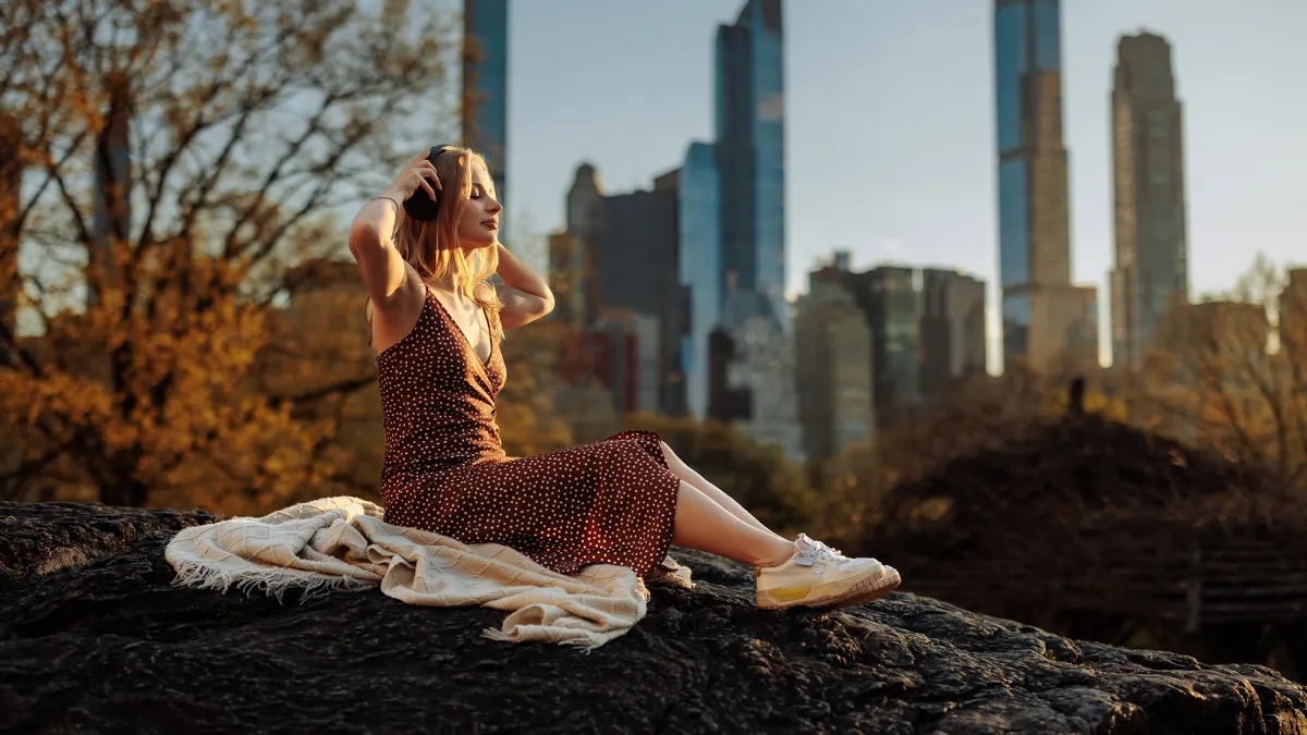 A woman sits serenely with a blanket, gazing at a city skyline from a vantage point, invoking a sense of peace despite the busy city backdrop