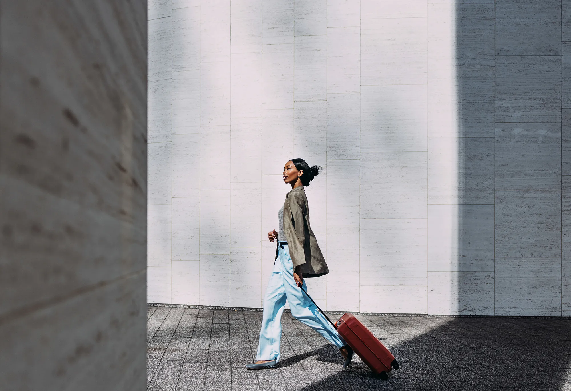 Confident woman walking with red suitcase against a modern urban wall. Ideal for travel, business, and lifestyle concepts.