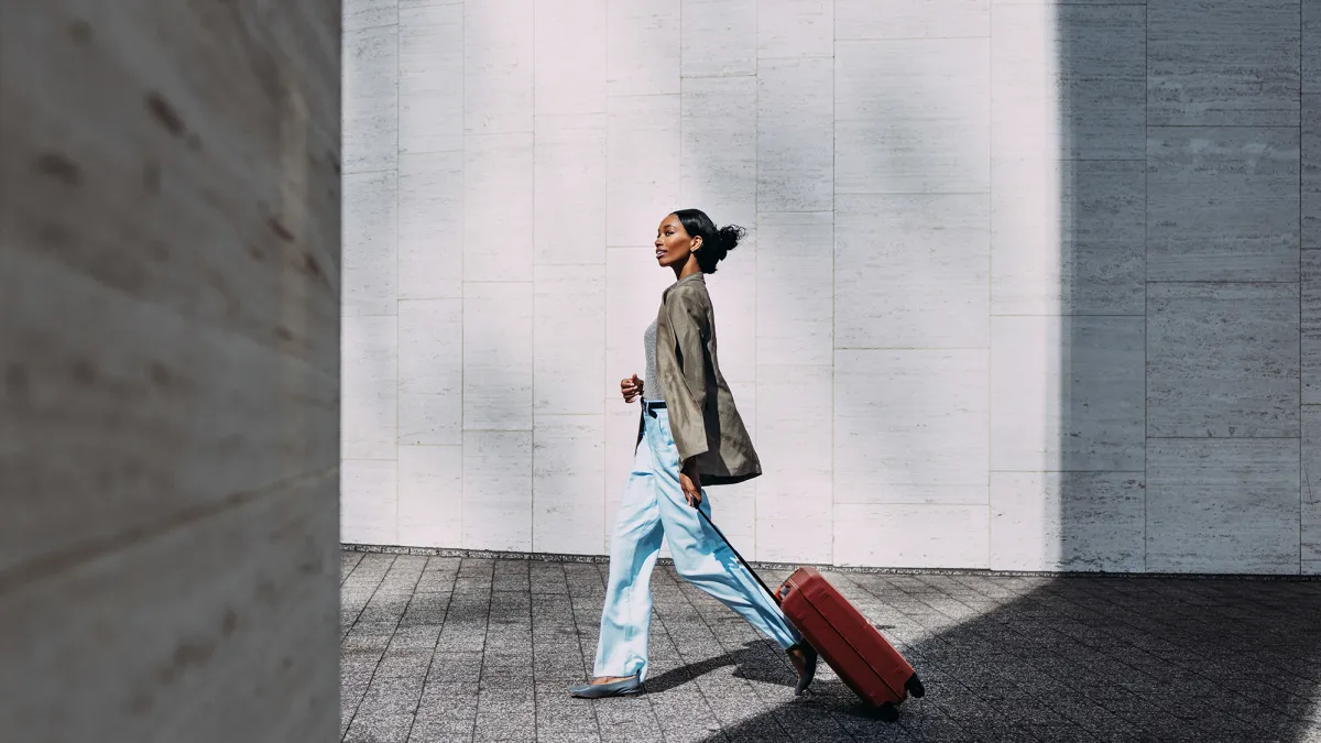Confident woman walking with red suitcase against a modern urban wall. Ideal for travel, business, and lifestyle concepts.