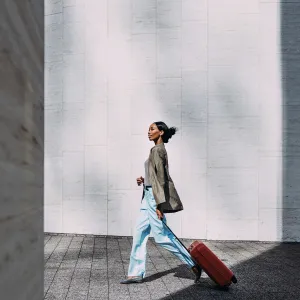 Confident woman walking with red suitcase against a modern urban wall. Ideal for travel, business, and lifestyle concepts.