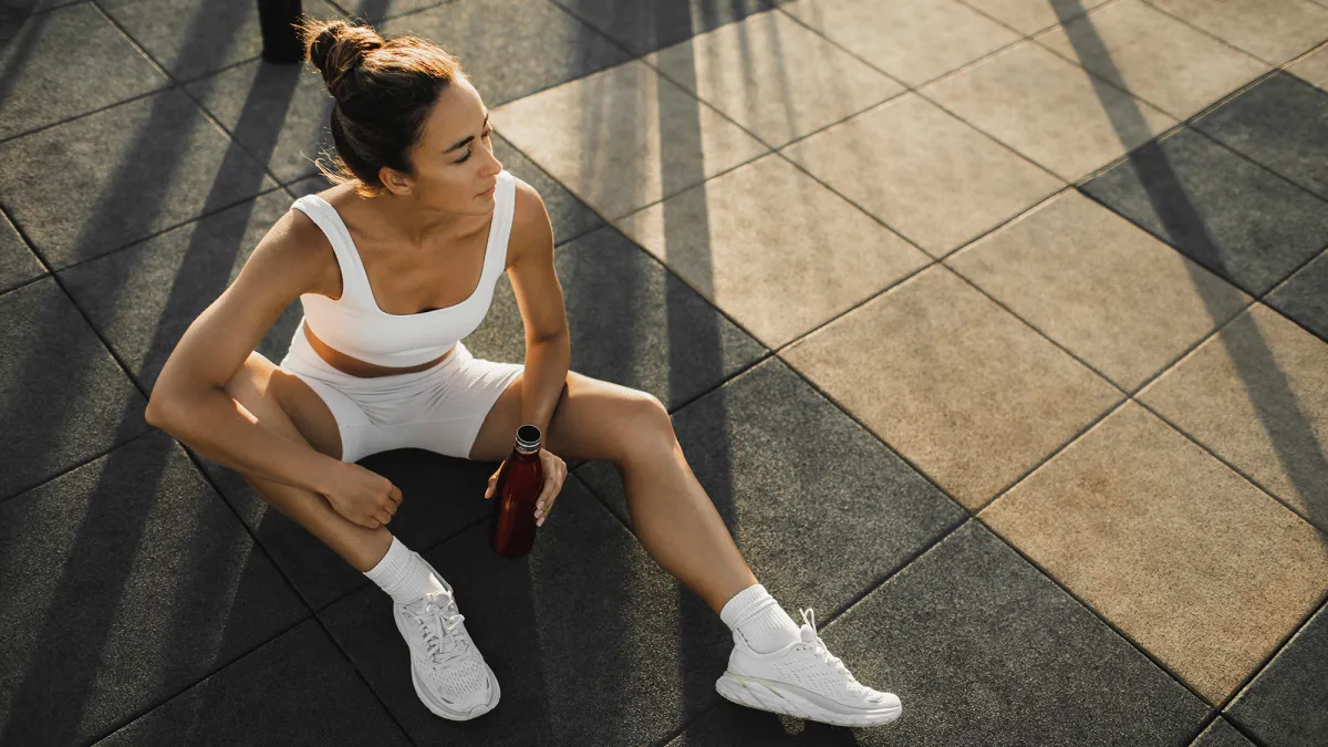 Sportive woman with slim tanned body relaxing after fitness training on outdoors sports ground. Taking a break to drink cold water and refresh. Sports hobby, healthy and active lifestyle concept.