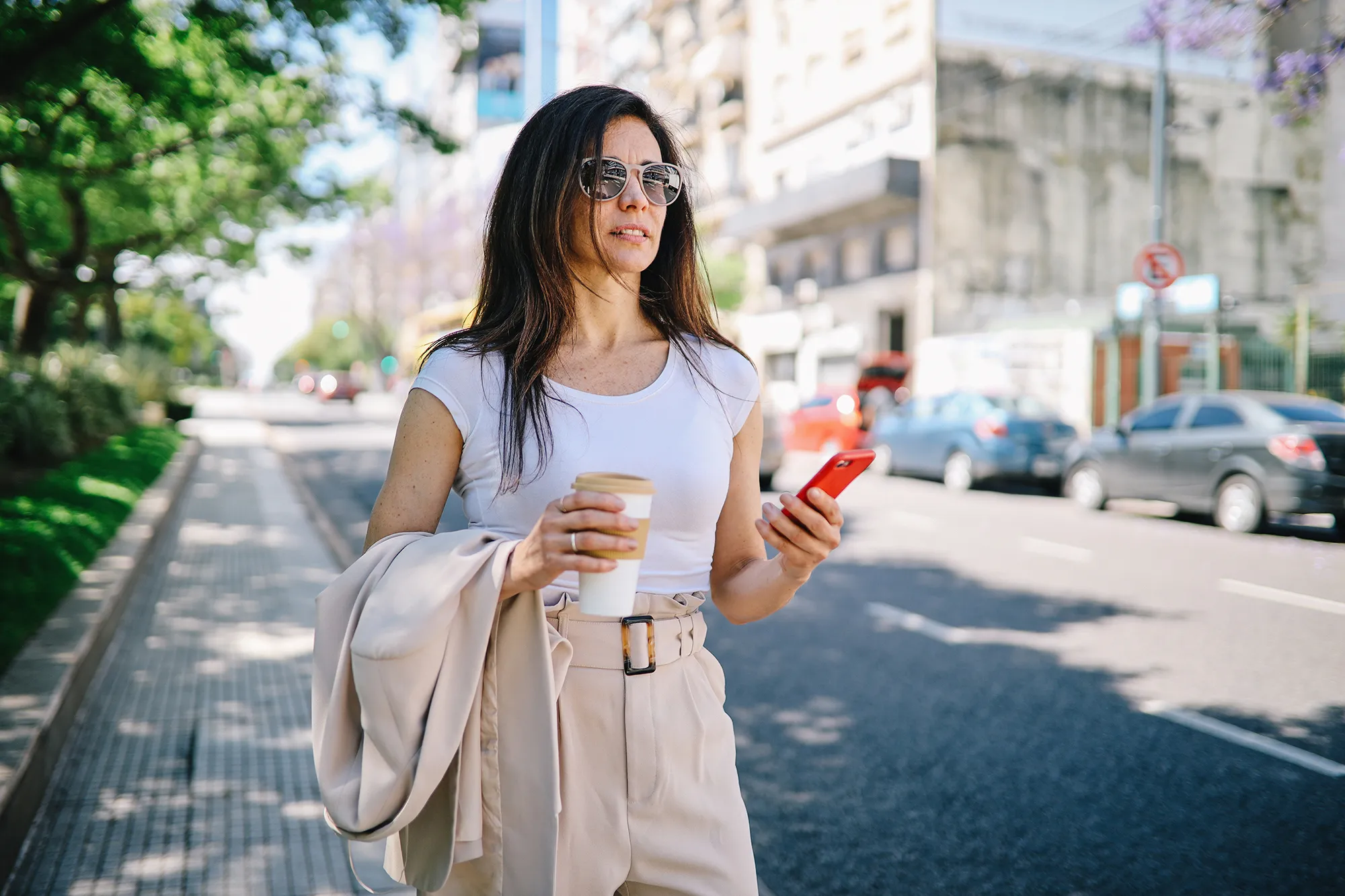 Fashionable woman walking on the street of Buenos Aires, looking for a taxi by using the mobile app on her smartphone.