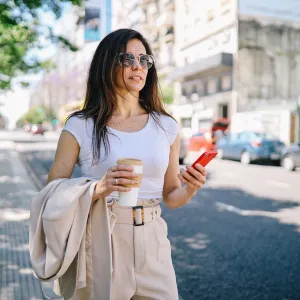 Fashionable woman walking on the street of Buenos Aires, looking for a taxi by using the mobile app on her smartphone.