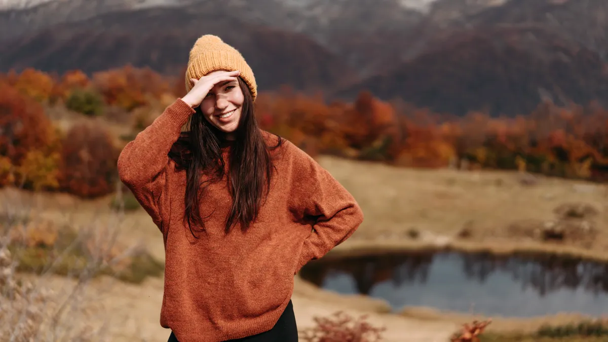 Cute young woman in a sweater and hat in the mountains by the lake in the autumn season.
