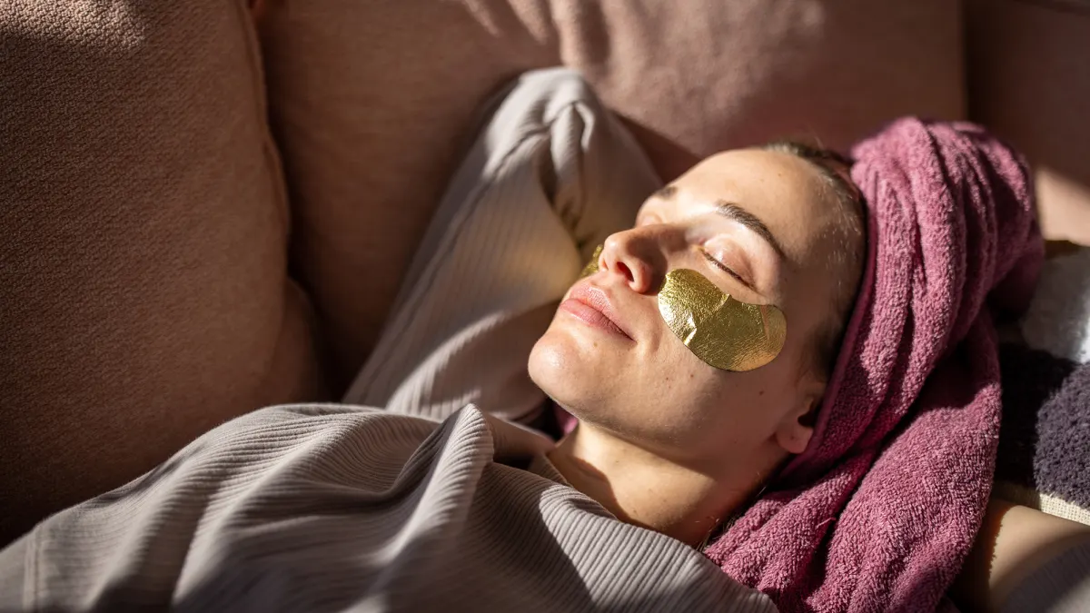 Young woman with eyes closed relaxing on sofa with towel wrapped around hair and gel patches under eyes.