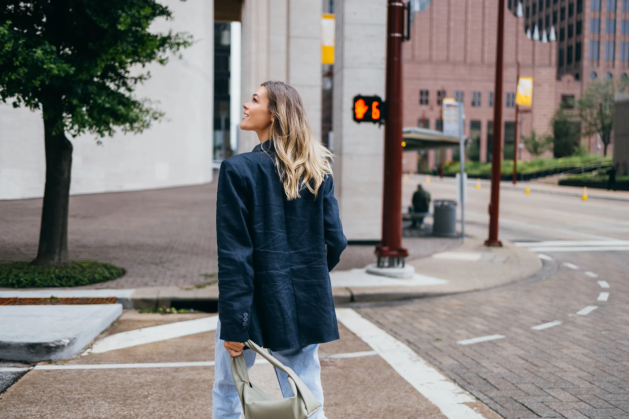 Rear view of a woman walking in an urban area, looking upward. She is dressed in casual attire with a handbag.