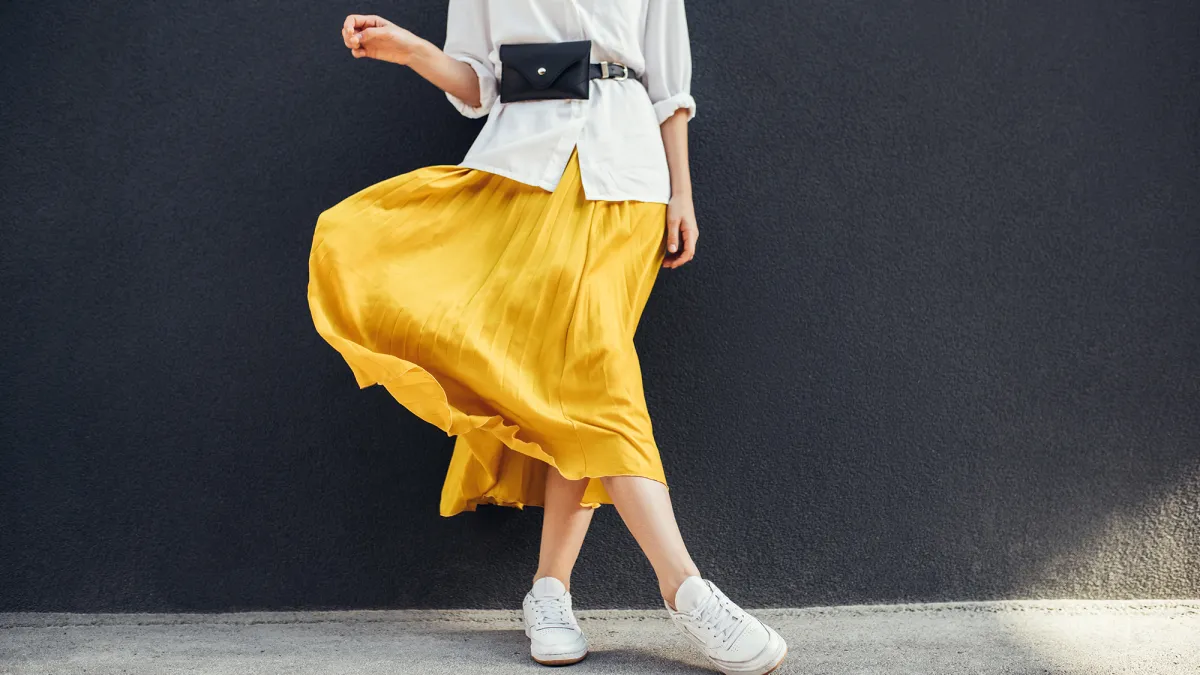 Horizontal cropped image of stylish slim woman in beautiful yellow skirt. Caucasian female fashion model standing over gray wall background outdoor with copy space.