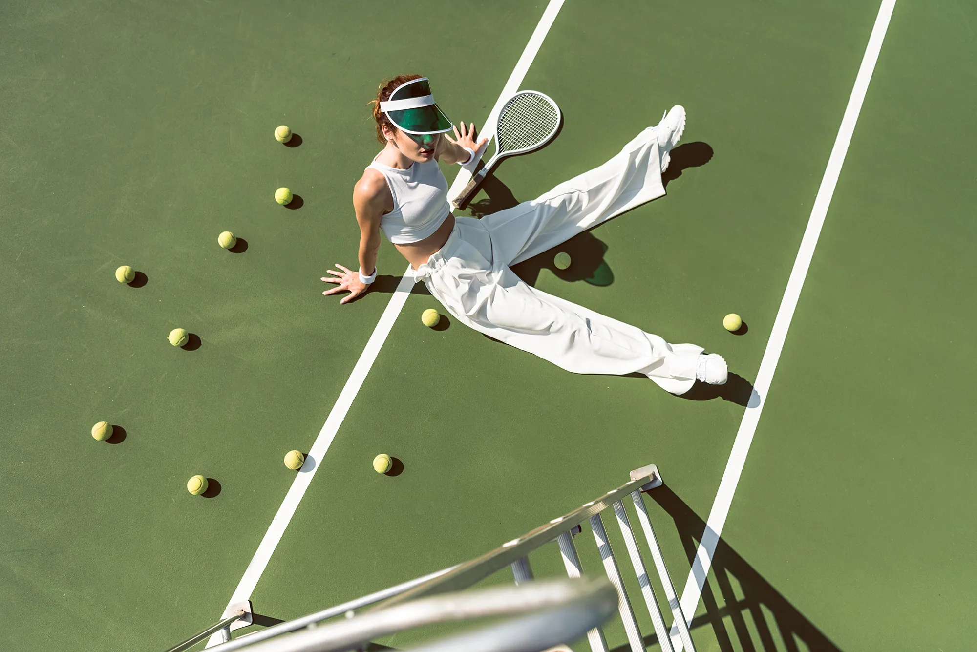 high angle view of beautiful woman in stylish white clothing sitting on tennis court with balls and racket around