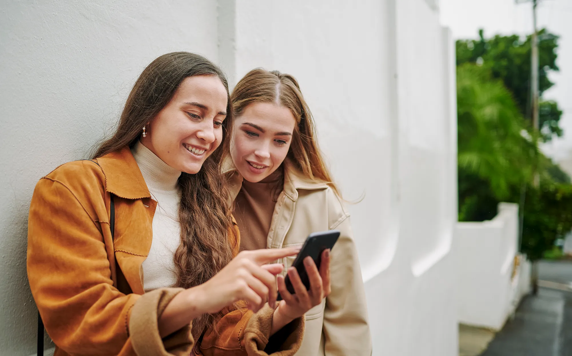 Two smiling young female friends swiping through an app on a smart phone while standing together on a city sidewalk