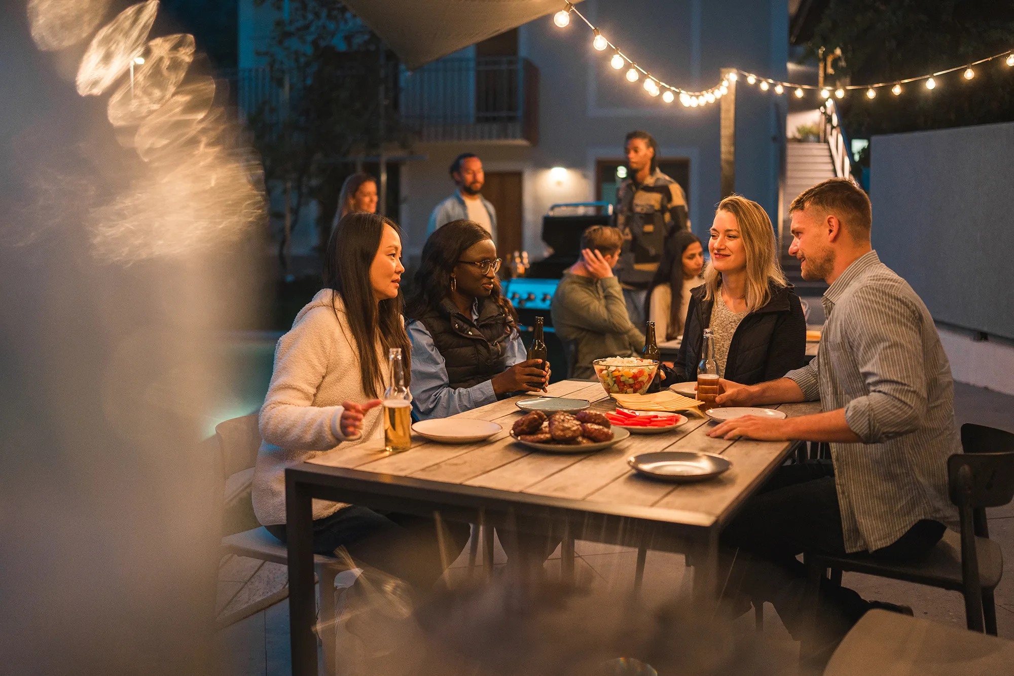 Friends gathered around a table, enjoying food and drinks at an evening outdoor party, with festive string lights above.