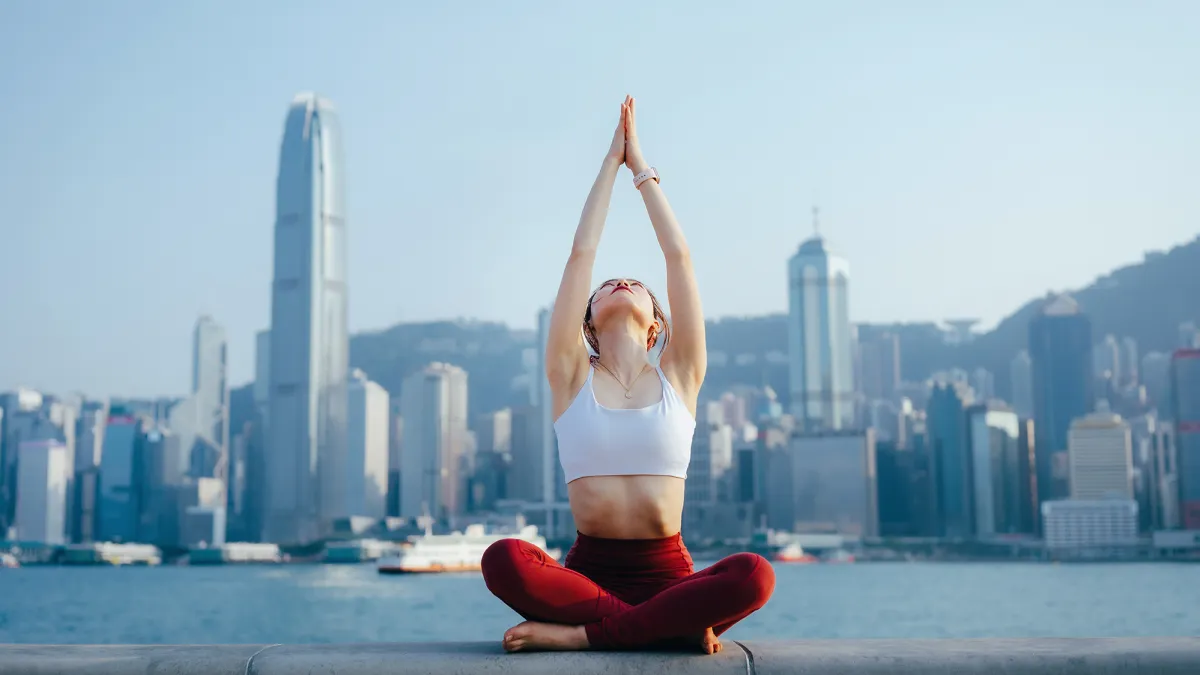Young woman meditating outdoors, practicing mindfulness yoga in the morning, against spectacular Hong Kong city skyline by the promenade of Victoria harbour.