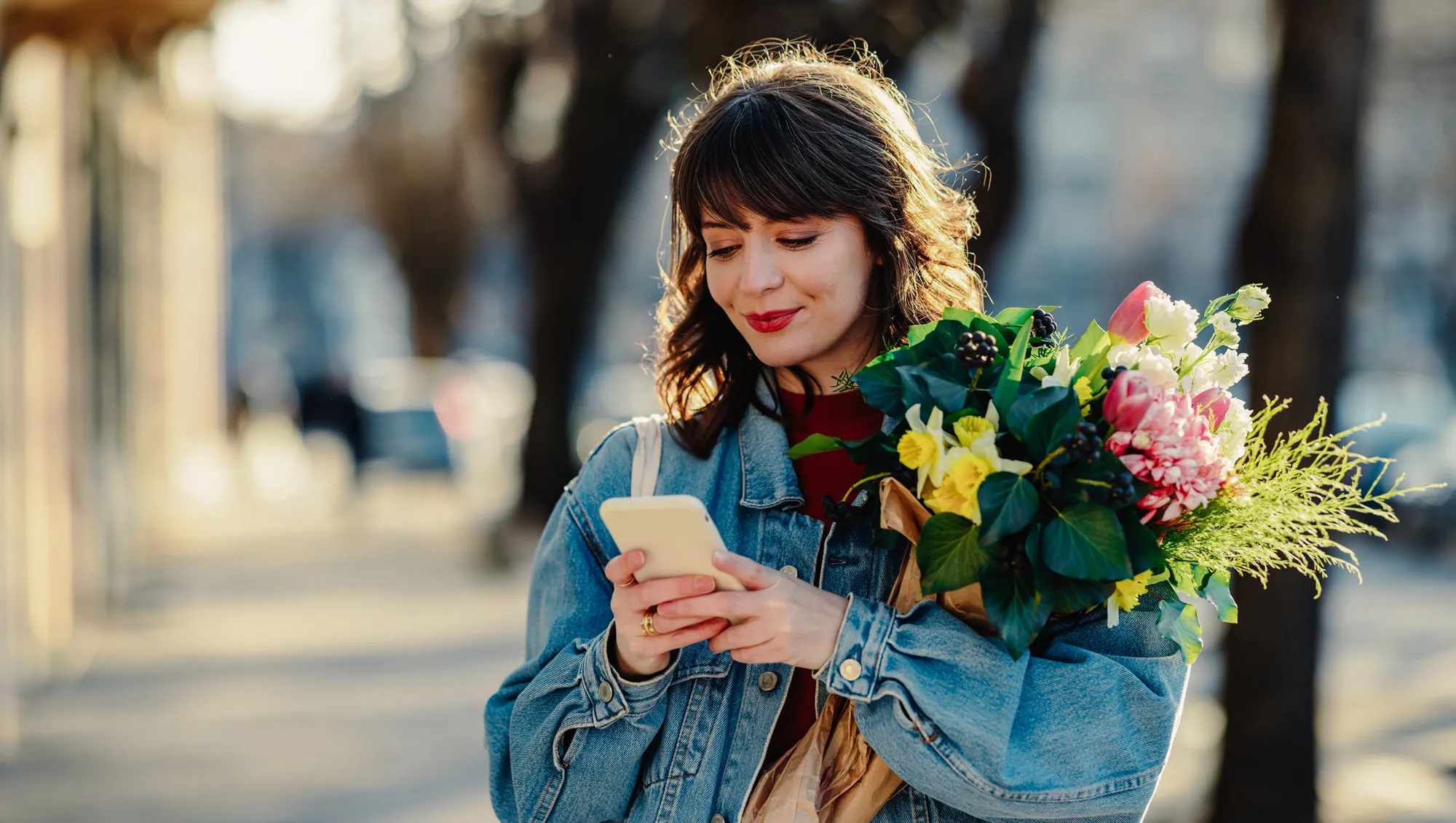 a woman holding a phone with a bouquet of flowers in a tree lined street