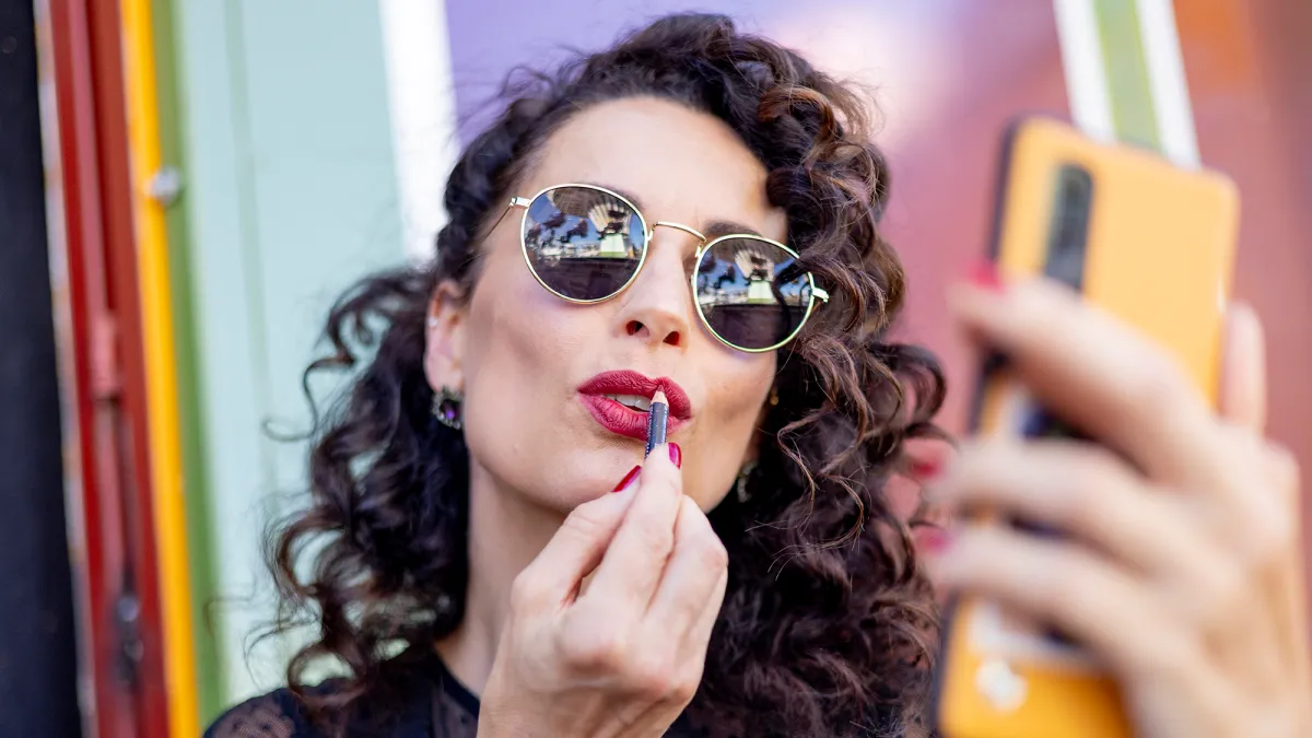 Woman with long dark curly hair and sunglasses applies red lipliner to her lips while using her smartphone as a mirror.
