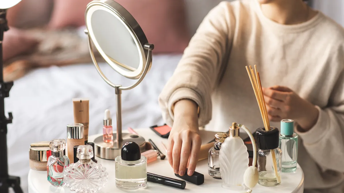 Cosmetics are laid out on a round white table. Beautiful little glass bottles and tubes with shiny lids, and some perfume. Woman is in the process of doing her make up.
