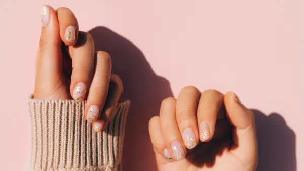 Glowing hands in sweater with knitted sleeves and with nude color manicure with gold particles on pink table. Trendy colors of the year