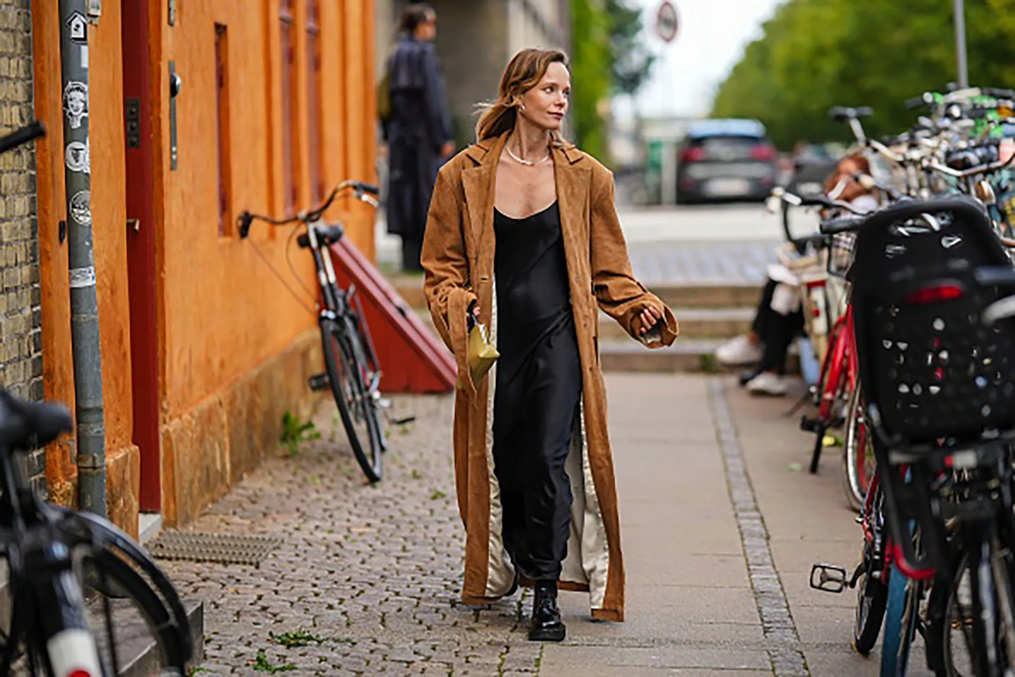 A guest wears silver earrings, a silver necklace, a black V-neck silk / satin long skirt, a dark brown suede long coat, black shiny varnished leather laces loafers, a pale yellow khaki shiny leather handbag , outside Remain, during the Copenhagen Fashion Week Spring/Summer 2024 on August 08, 2023 in Copenhagen, Denmark.