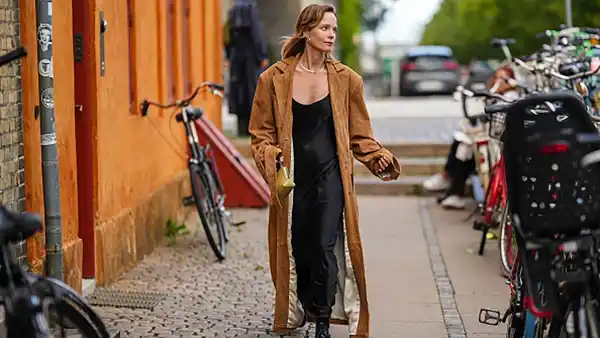 A guest wears silver earrings, a silver necklace, a black V-neck silk / satin long skirt, a dark brown suede long coat, black shiny varnished leather laces loafers, a pale yellow khaki shiny leather handbag , outside Remain, during the Copenhagen Fashion Week Spring/Summer 2024 on August 08, 2023 in Copenhagen, Denmark.