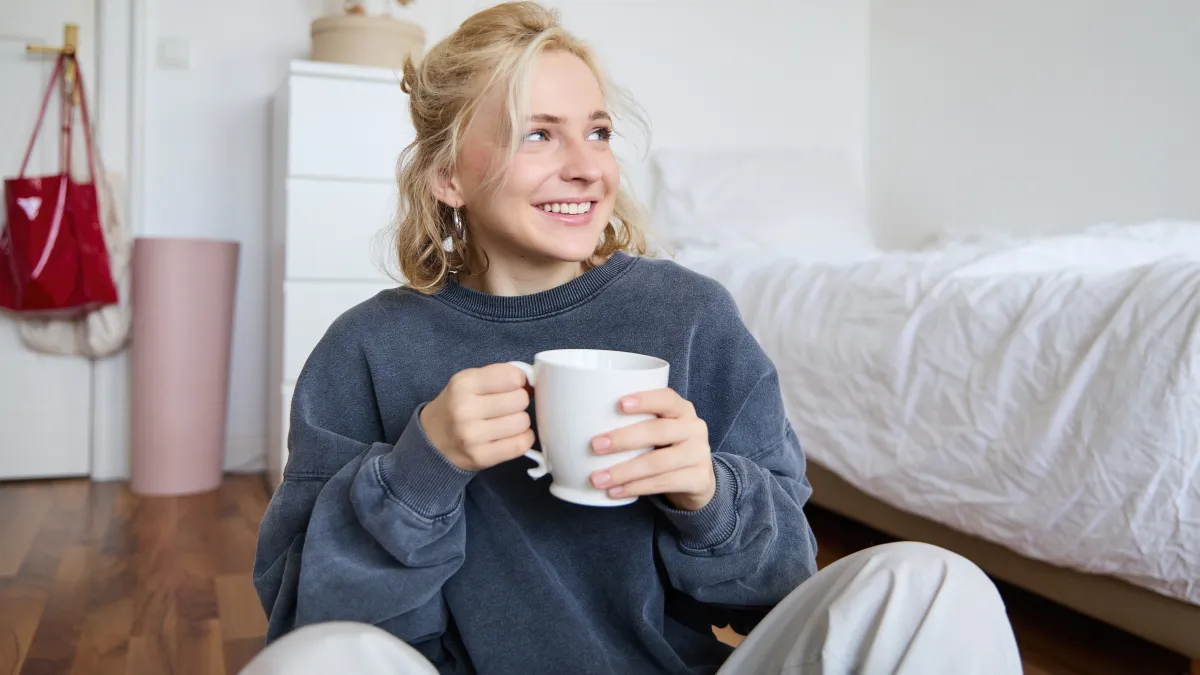Lifestyle portrait of young woman sitting on bedroom floor with cup of tea, drinking from big white mug and looking aside, smiling happily.