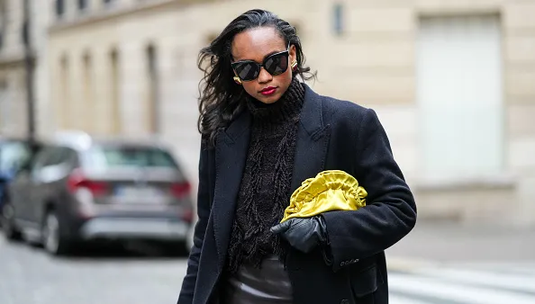 PARIS, FRANCE - OCTOBER 18: Emilie Joseph wears oversized double breasted wool extra long coat from COS, chocolate wool blended fringed turtleneck jumper from COS, lack leather long gloves, Black sunglasses from Gentle Monster, a satin yellow clutch/pouch bag, during a street style fashion photo session, on October 18, 2024 in Paris, France. (Photo by Edward Berthelot/Getty Images)