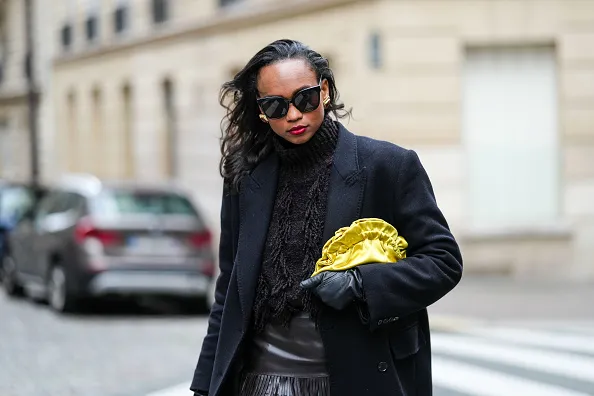PARIS, FRANCE - OCTOBER 18: Emilie Joseph wears oversized double breasted wool extra long coat from COS, chocolate wool blended fringed turtleneck jumper from COS, lack leather long gloves, Black sunglasses from Gentle Monster, a satin yellow clutch/pouch bag, during a street style fashion photo session, on October 18, 2024 in Paris, France. (Photo by Edward Berthelot/Getty Images)