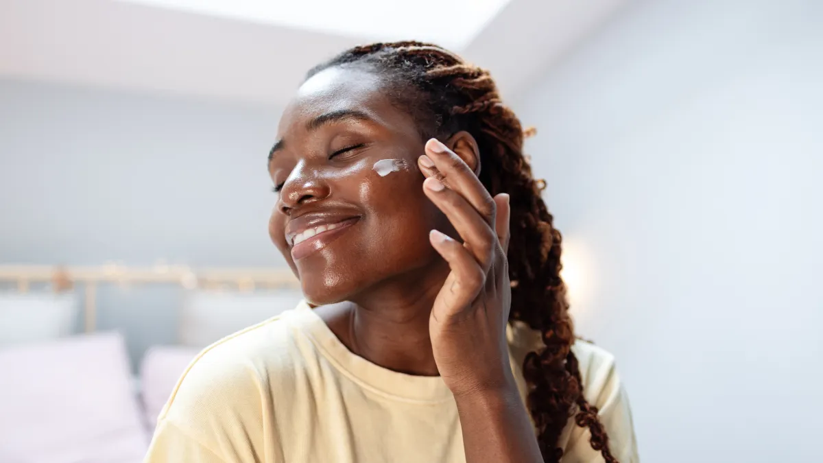 A close-up of a young African American woman cheerfully smiling, while applying beauty cream with her hand on her face.