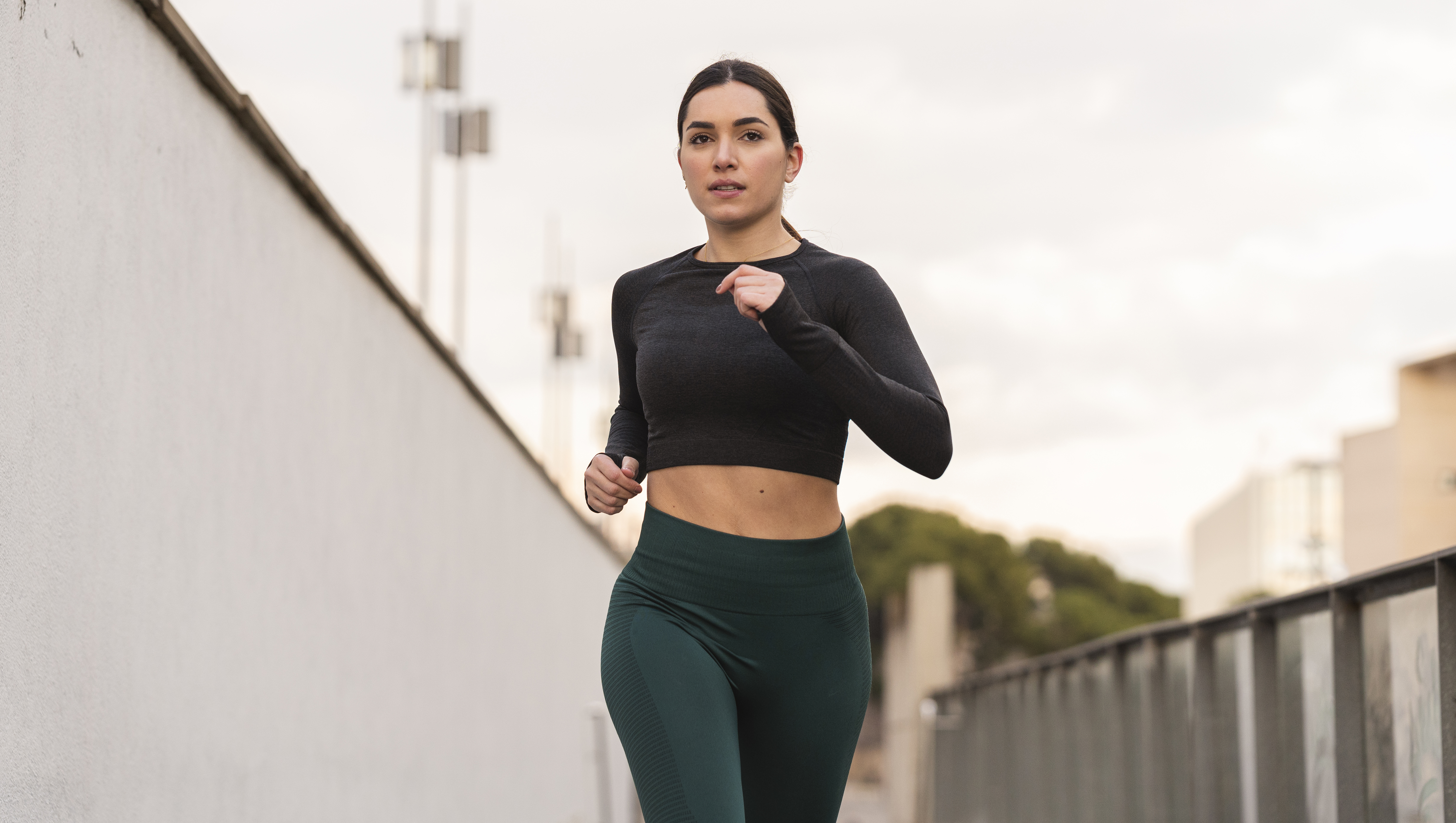 Medium shot of a young latina female athlete running towards the camera during her daily routine. She is wearing a dark grey long sleeve t-shirt and green leggings. With copy space